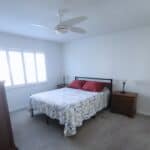 Bedroom with large window and ceiling fan, featuring a comfortable bed with red pillows and wooden n.
